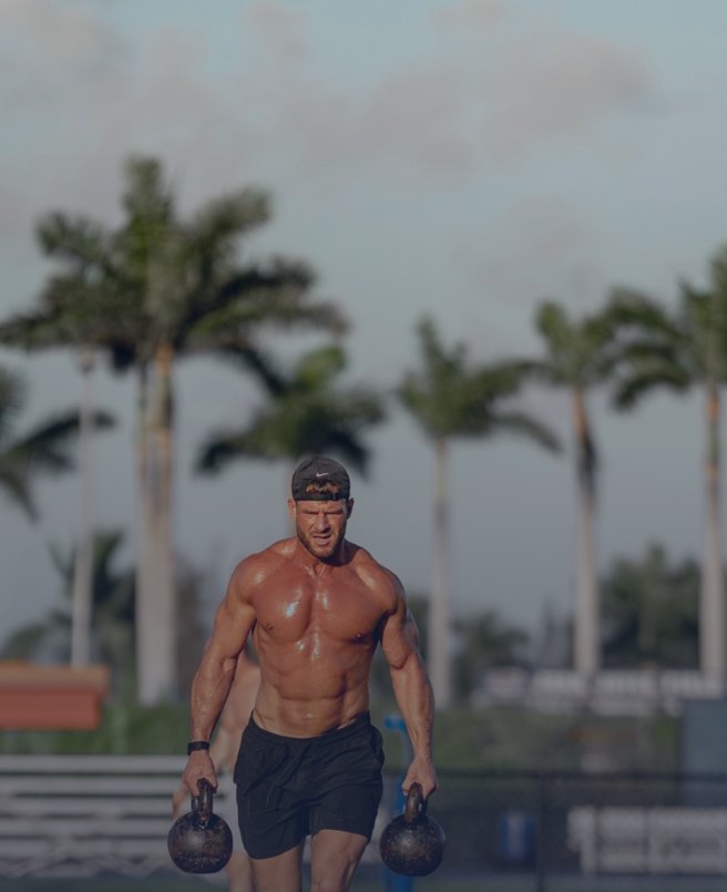 Zach White, founder of BioKingdom Health, carrying two kettlebells outdoors beneath palm trees